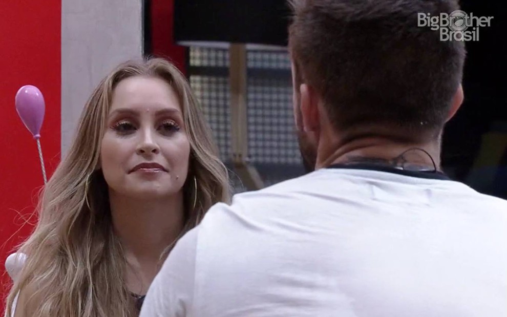 Carla olha para Arthur; ela está com o cabelo solto, e ele usa camiseta branca