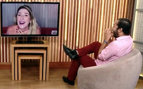 Dani está aparecendo em uma televisão, ela está com a boca aberta e as mãos no pescoço; Gil olha para a tela, usa camiseta rosa, calça vermelha e sapato preto