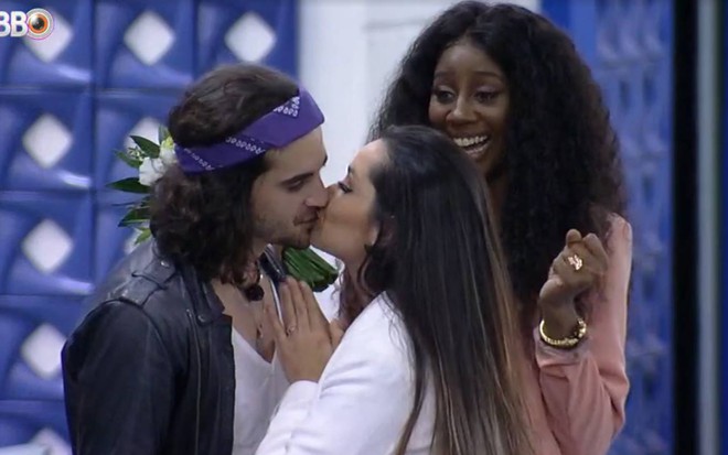 Fiuk e Juliette estão se beijando, ele usa jaqueta preta e camiseta branca, ela usa casaco branco e está de cabelo solto; Camilla usa casaco rosa e sorri olhando para os dois