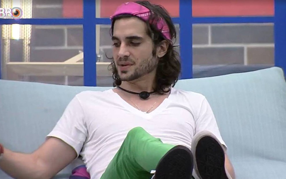 Fiuk está sentado em um sofá azul, usa calça verde, camiseta branca e lenço rosa no cabelo