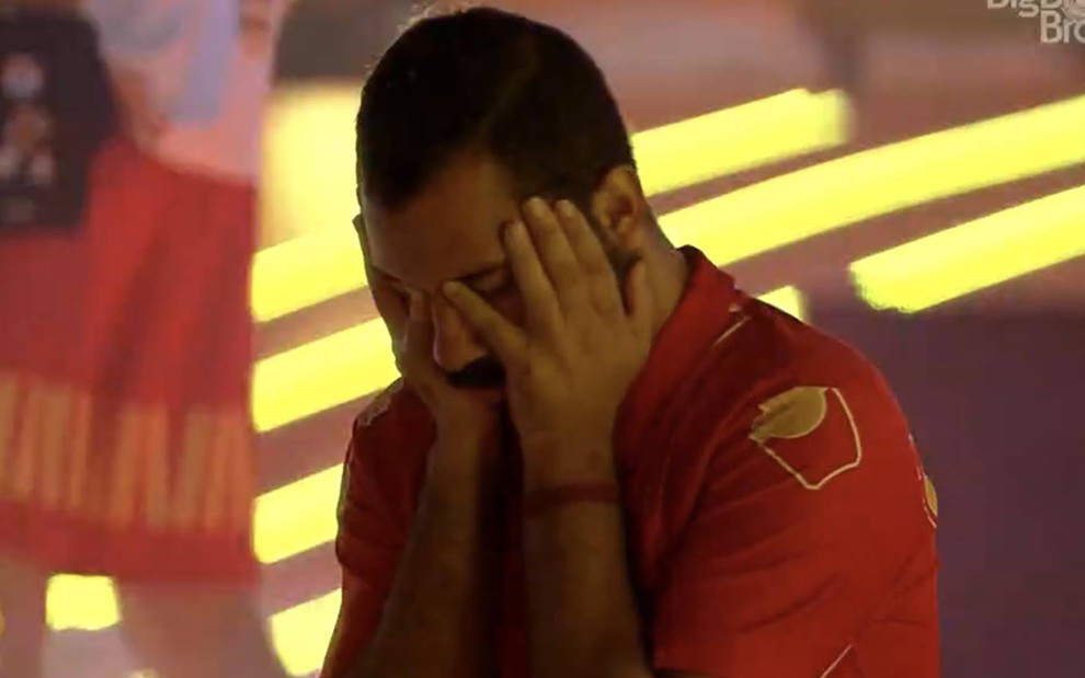 Gilberto Nogueira olha para baixo, usa camiseta vermelha e está com as duas mãos no rosto