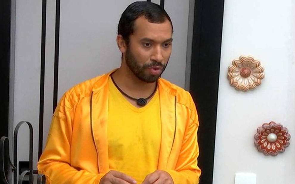 Gilberto olha para baixo, usa camiseta amarela e jaqueta amarela; ele está em pé na frente da porta branca