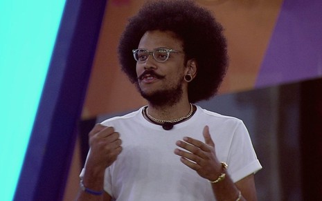 João Luiz Pedrosa na área externa do BBB21 com mãos gesticulando, expressão séria, camiseta branca