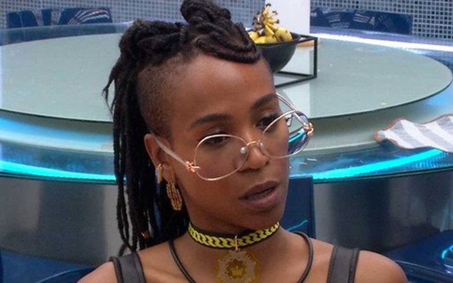 Karol olha para o lado; ela usa blusa de alça preta, colar amarelo e óculos de grau