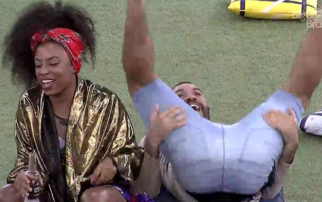 Sentada no chão, Lumena ri, está com o cabelo preso com um lenço vermelho e casaco dourado; Gilberto está jogado no chão com as pernas pra cima; ele usa bermuda jeans