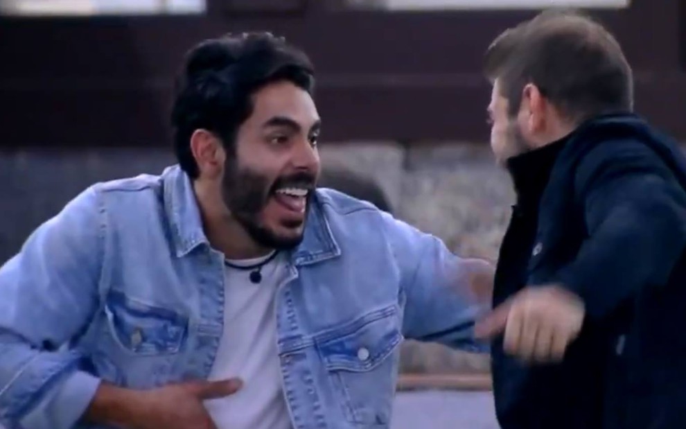 Rodolffo sorri, olha para Caio e usa jaqueta jeans com camiseta branca; Caio está de costas e usa jaqueta preta