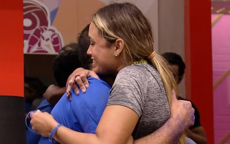 Sarah está abraçada com Gilberto; ela veste camiseta cinza e está com o cabelo preso; Gilberto está com camiseta azul