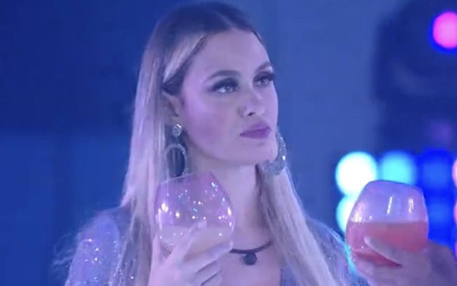 Sarah olha para o lado, está com o cabelo solto e usa vestido prata