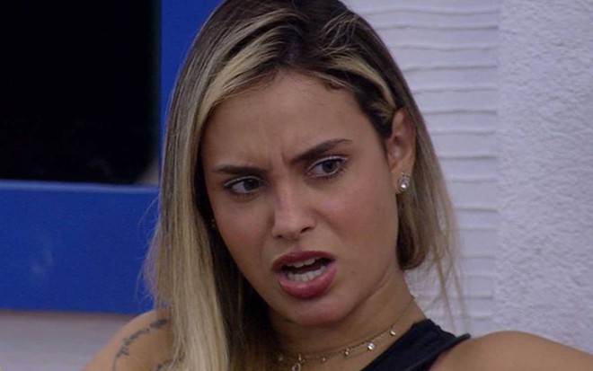 Sarah olha para o lado, usa regata preta e está com o cabelo solto; a sister está com a boca aberta