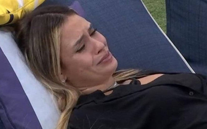 Sarah está deitada em uma cadeira azul, está chorando e usa blusa preta