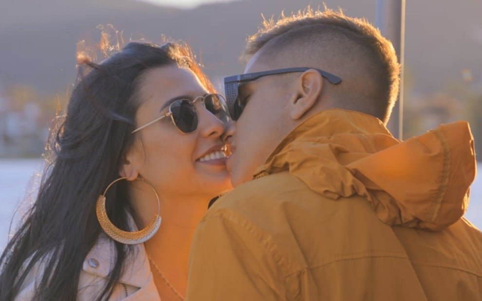 Imagem de uma mulher de cabelo preto e um capaz de blusa amarela se beijando com o mar ao fundo