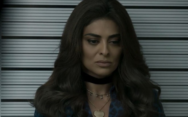 A atriz Juliana Paes (Bibi) está com expressão séria e com a cabeça baixa; ela aparece em frente a uma parede de reconhecimento de suspeito na delegacia