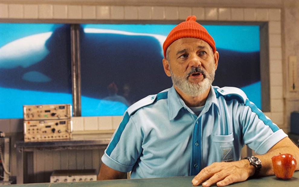Bill Murray como Steve Zissou em A Vida Marinha Com Steve Zissou, vestido com camisa azul clara e com touca vermelha na cabeça, ele está sentado ao lado de uma maçã mordida com expressão de dúvida