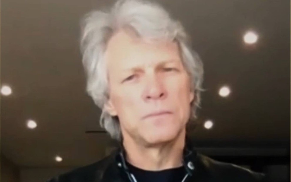 Jon Bon Jovi olha para a câmera, usa camiseta preta e jaqueta preta