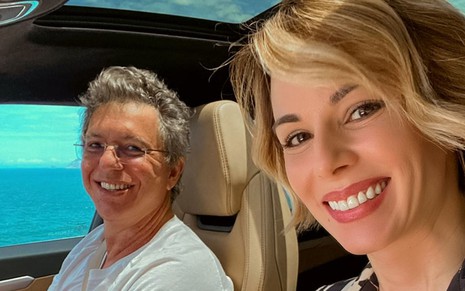 Boninho e Ana estão dentro do carro, ela sorri para a foto; ele também sorri e usa camiseta branca