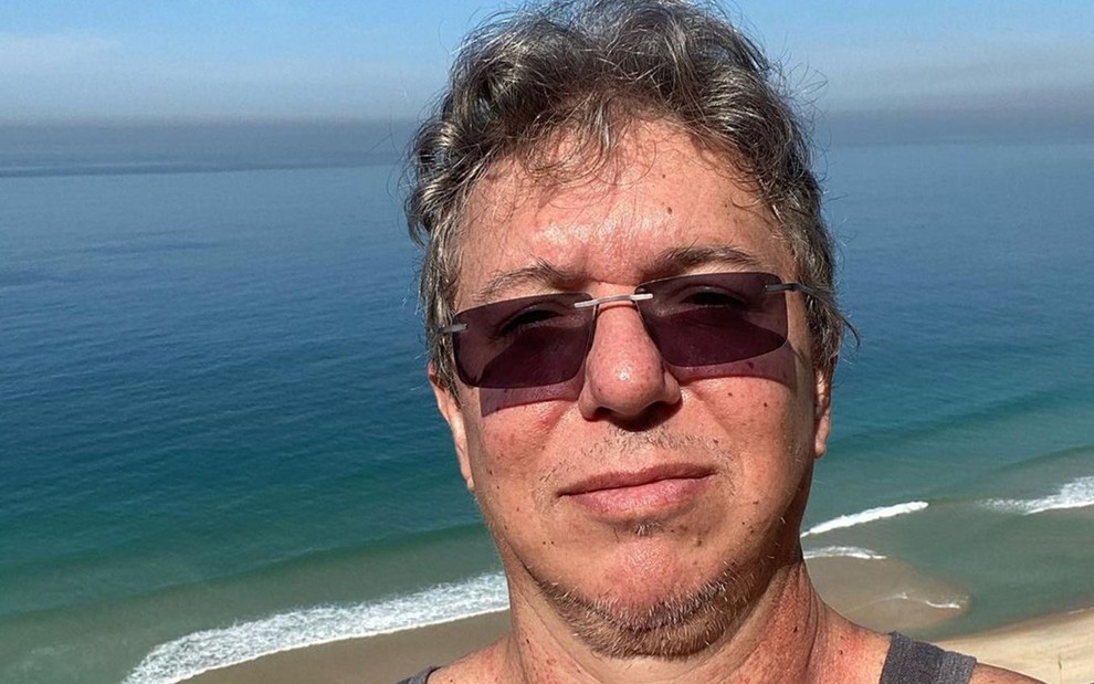 Boninho está tirando uma selfie; ele usa óculos de sol e usa regata cinza; ele está de costas para o mar