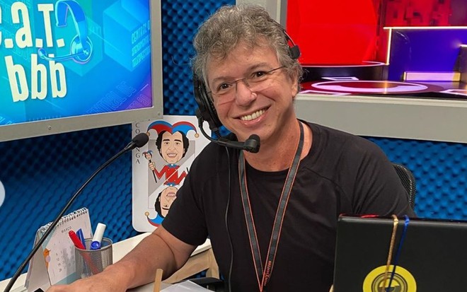 Boninho olha para frente, sorri e usa óculos de grau; ele veste uma camiseta preta e está com um fone na cabeça e microfone na boca