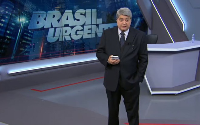 O apresentador José Luiz Datena de terno preto, de pé, no estúdio do Brasil Urgente, na Band