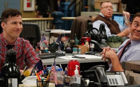 Andy Samberg sorri ao olhar para Joe Lo Truglio, com Joel McKinnon Miller ao fundo, na série Brooklyn Nine-Nine 