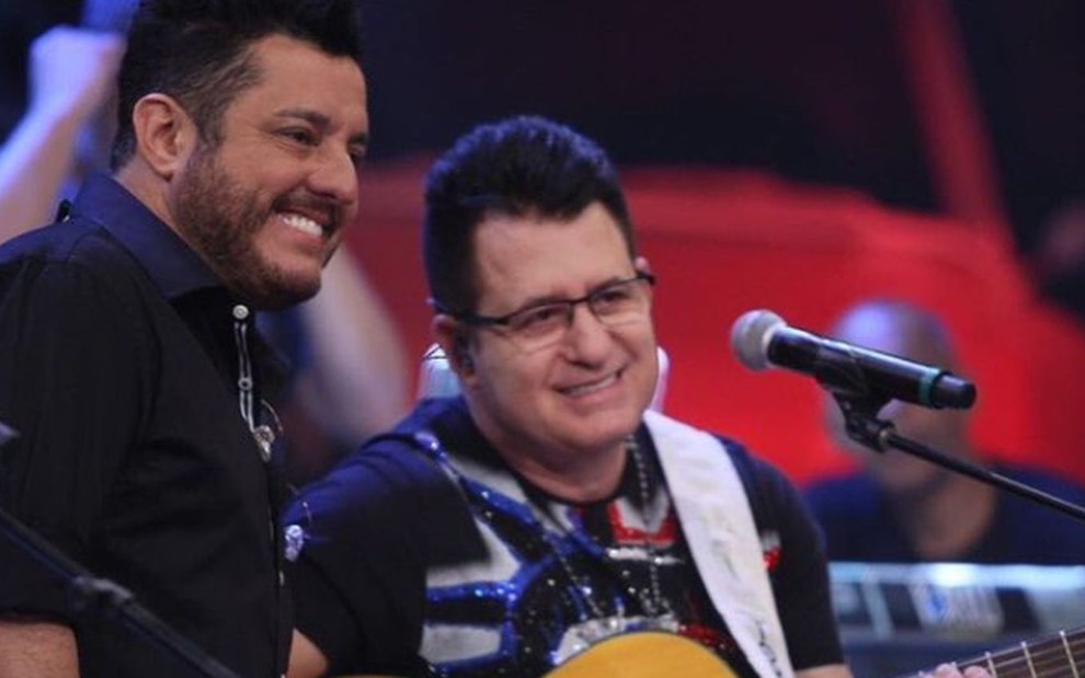 Bruno e Marrone sorridentes, abraçados; Bruno em pé, de camisa preta; Marrone sentado, com violão
