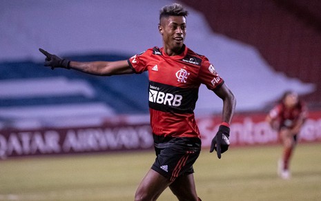 Bruno Henrique com o uniforme vermelho e preto do Flamengo apontando para um lado e olhando para o outro