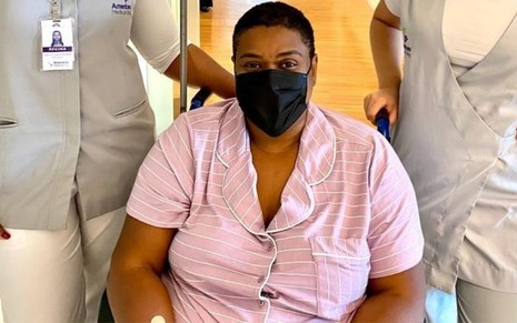 Cacau Protásio na cadeira de rodas entre duas enfermeiras no hospital