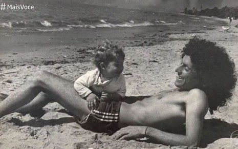 Caco Barcellos mais jovem, com visual de hippie, cabelo encaracolado comprido, mais magro, na praia