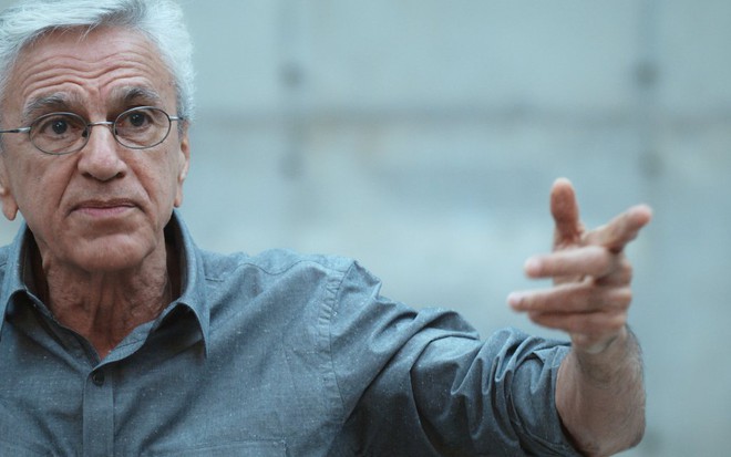 Caetano Veloso com expressão séria em trecho do documentário Narciso em Férias, com fundo cinza e camisa cinza