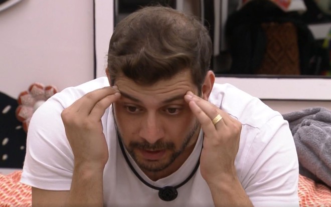 Caio Afiune, participante do BBB21, usa camisa branca enquanto leva as mãos à cabeça