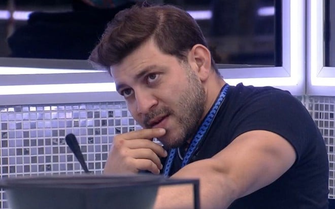 Caio Afiune olha para o horizonte com a mão no queixo na cozinha do BBB21