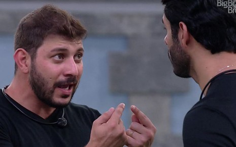 Caio conversa com Rodolfo, usa camiseta preta e está de boca aberta; Rodolffo está de costas e usa camiseta preta