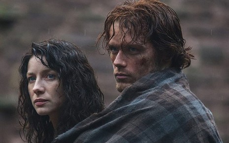 Sujos e cobertos com uma manta azul xadrez, Claire (Caitriona Balfe) e Jamie (Sam Heughan) estão sérios em cena de Outlander