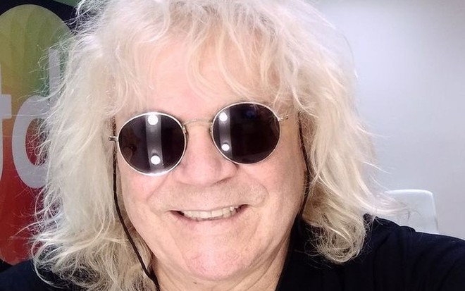 O cantor Ovelha com o cabelo loiro cacheado, camiseta preta e óculos escuros