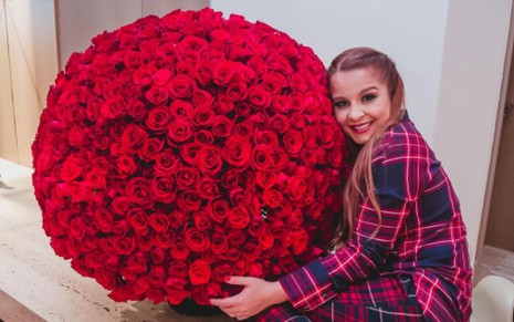 A cantora Maiara de pijama xadrez, vermelho e azul, abraçada a um buquê de rosas gigante