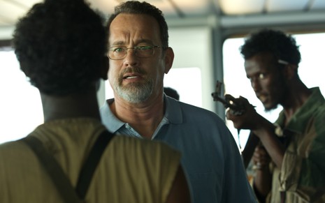 Tom Hanks ao centro olha para frente, onde tem um homem parado; atrás dele, tem um homem com uma arma apontada