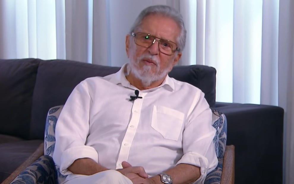 O apresentador Carlos Alberto de Nóbrega usa camisa branca e óculos sentado em um sofá escuro