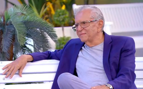 Carlos Alberto da Nóbrega está sentado no banco do estúdio do programa A Praça É Nossa. Ele está vestido um paletó azul por baixo de uma camiseta azul