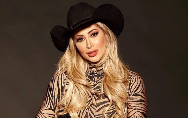 Carol Narizinho posa em um fundo preto; está usando uma blusa com estampa de zebra de tom marrom claro. O cabelo loiro está solto e ela usa um chapéu preto