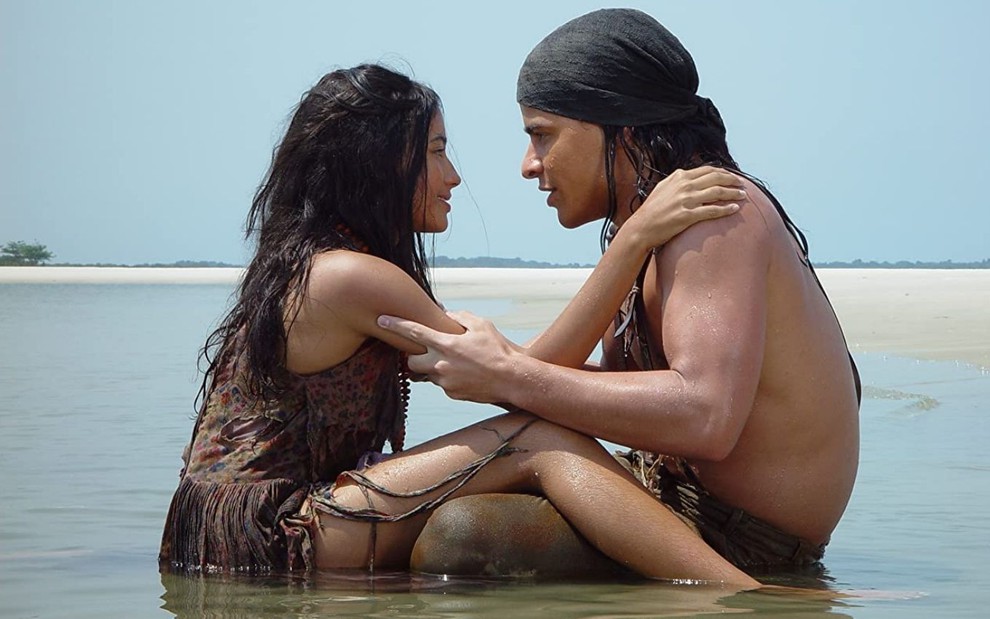 Sentados na beira do mar, Carolina Oliveira e Thiago Martins se encaram em cena do filme Encantados (2017)