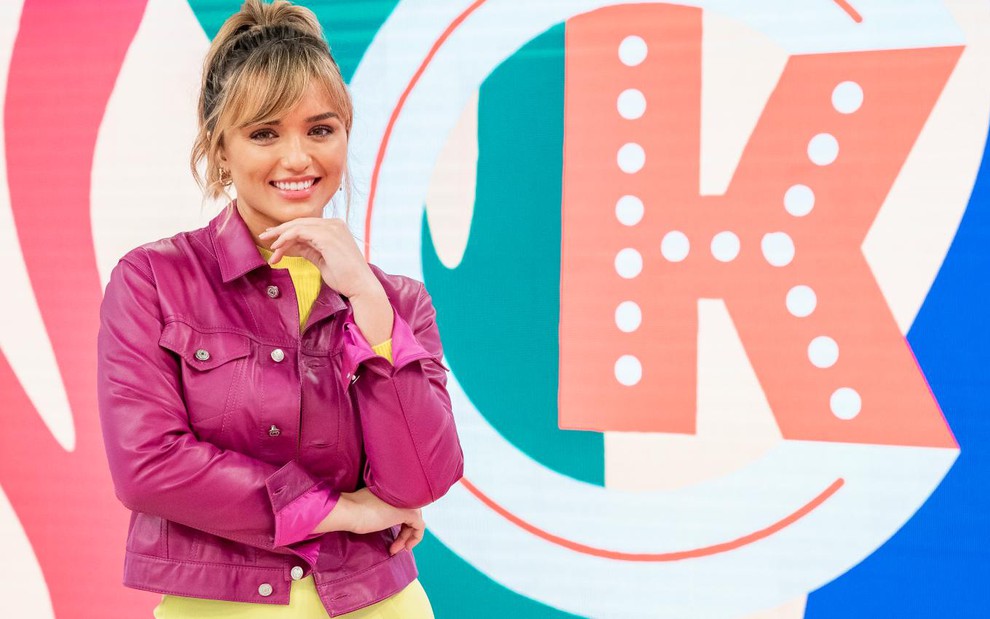 Rafa Kalimann sorri, com a mão esquerda no queixo, no cenário do programa Casa Kalimann; uma letra K aparece ao lado dela