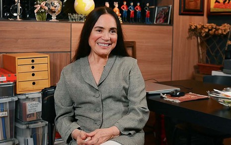 Regina Duarte sorri e está sentada em um escritório