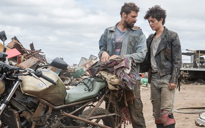 Ao lado de uma moto, Cauã Reymond como Ara e Sophie Charlotte como Severina em Reza a Lenda, ambos com roupas batidas e semblante de preocupação.