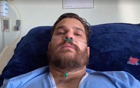 Cantor Cauan com roupa de hospital azul, deitado na maca, com respirador no nariz