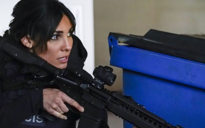 A atriz Daniela Ruah, com uniforme tático, segura arma em cena de ação de NCIS: Los Angeles