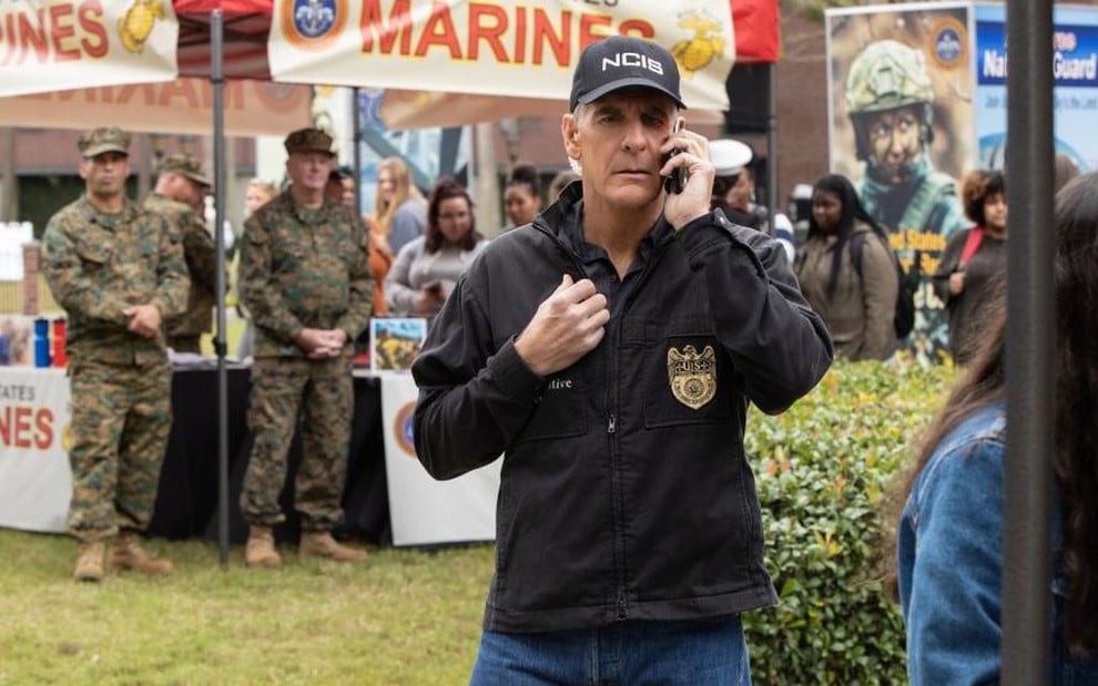 Com jaqueta de polícia e um boné escrito NCIS, o ator Scott Bakula fala ao celular em cena da série NCIS: New Orleans