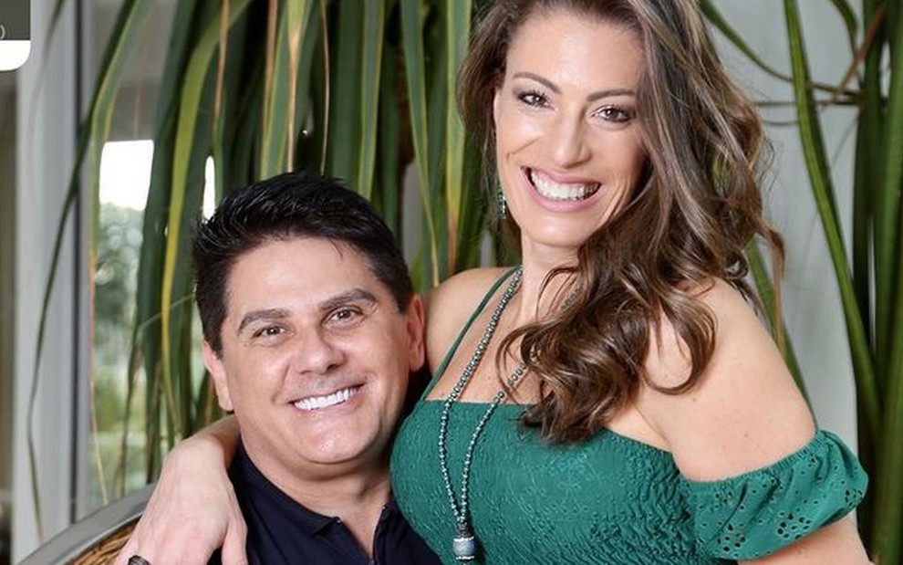 Cesar Filho sorridente, com camiseta azul marinho, e a mulher dele, Elaine Mickely, de vestido verde sentada em seu colo