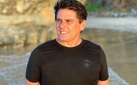 O apresentador Cesar Filho numa praia, com camiseta preta e cabelo molhado, olha patra a esquerda