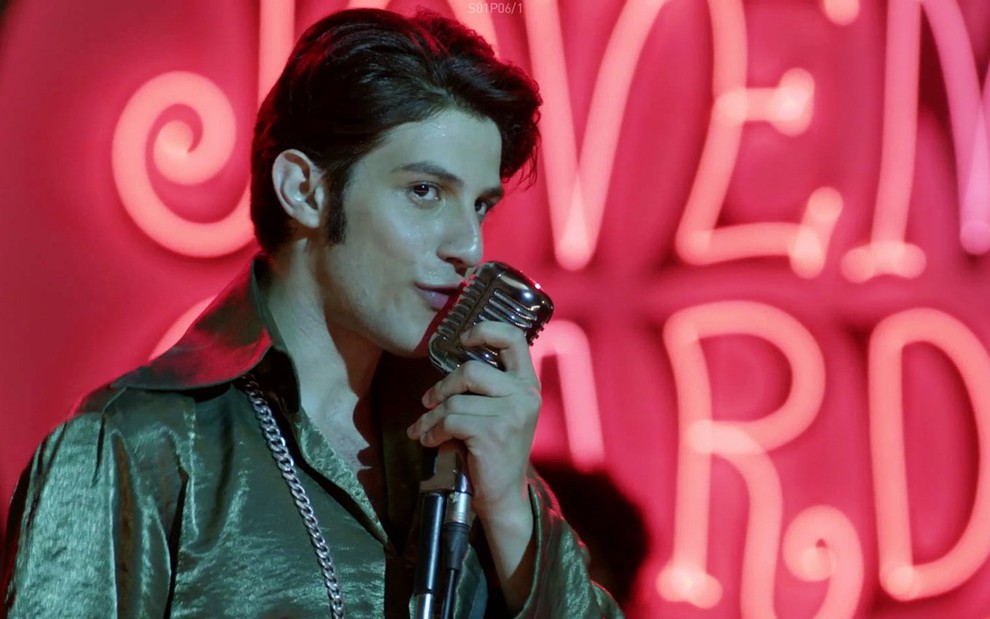 Chay Suede vestindo uma jaqueta, cantando perto de um microfone, na frente de uma parede rosa escrita Jovem Guarda
