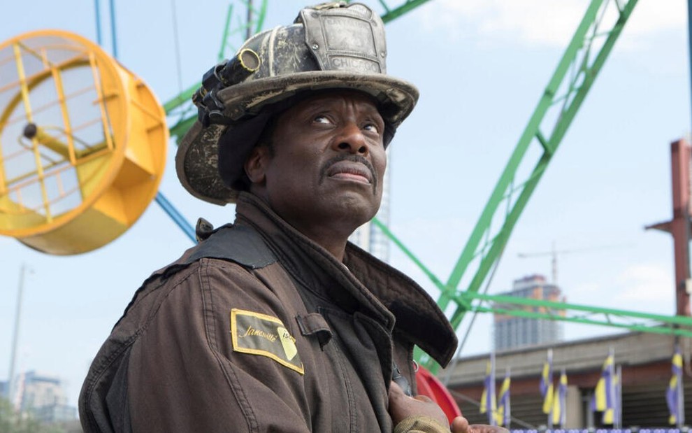 Em um parque de diversões, o ator Eamonn Walker aparece com semblante série, vestindo um uniforme de bombeiro