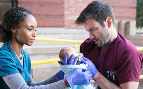 Na pele de uma enfermeira, vestindo azul, Yaya da Costa entrega um bebê para Colin Donnell, que é um médico em Chciago Med